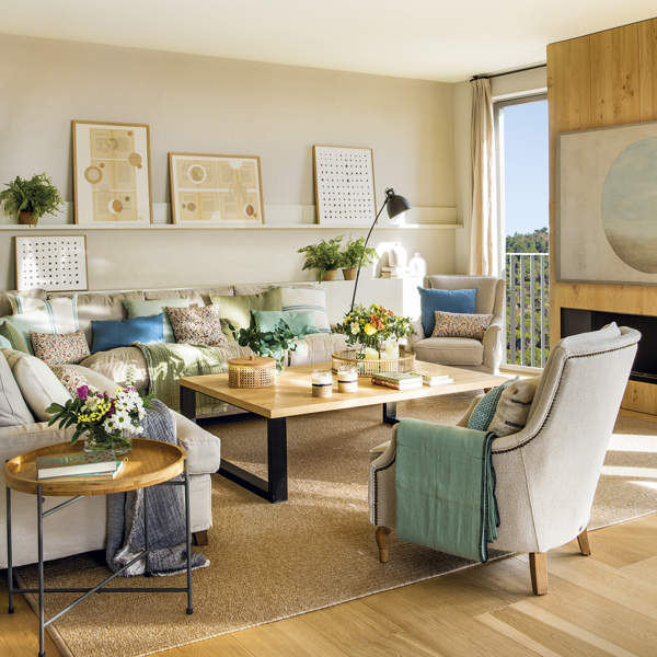 salón con chimenea y sofá gris esquinero con cojines azul, verde y estampados, mesa de centro de madera con patas de metal. Cuadros en estantería en la pared